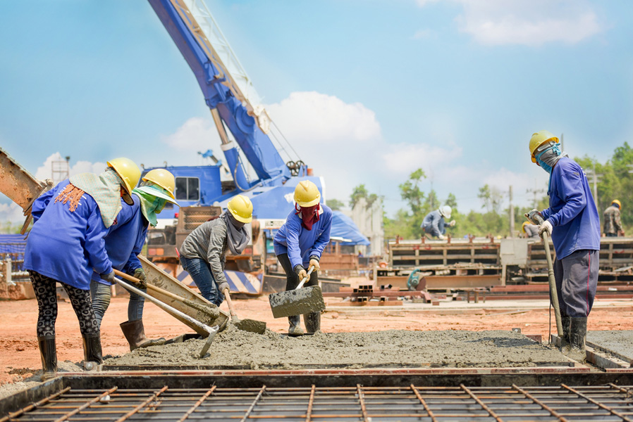 The image depicts a construction site where workers in safety gear are pouring and spreading concrete over reinforcement bars, with a crane in the background.