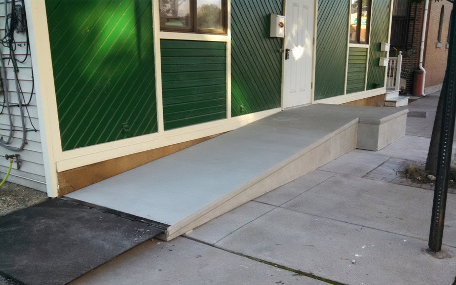 The image shows a concrete ramp leading to a building entrance with green siding and white trim. There's a black rubber mat at the base of the ramp.