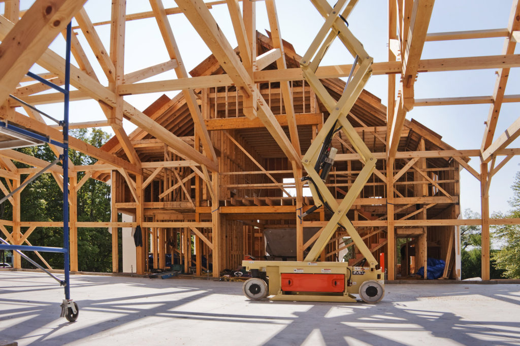 The image shows the construction of a wooden residential building with exposed beams and framework. A scissor lift is visible in the foreground.