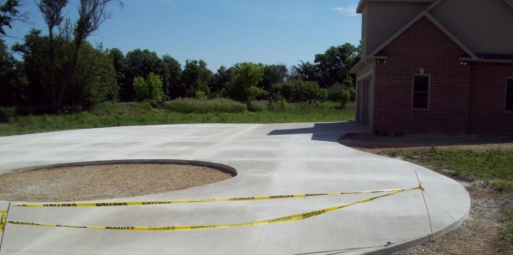 Newly poured circular concrete driveway with caution tape around, next to a brick house with trees.