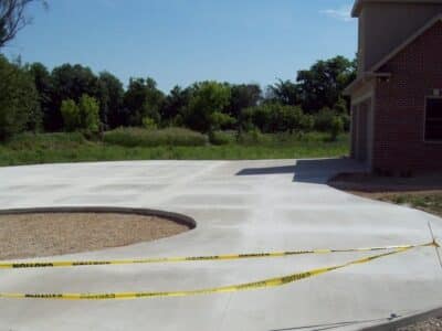 Newly poured circular concrete driveway with caution tape around, next to a brick house with trees.