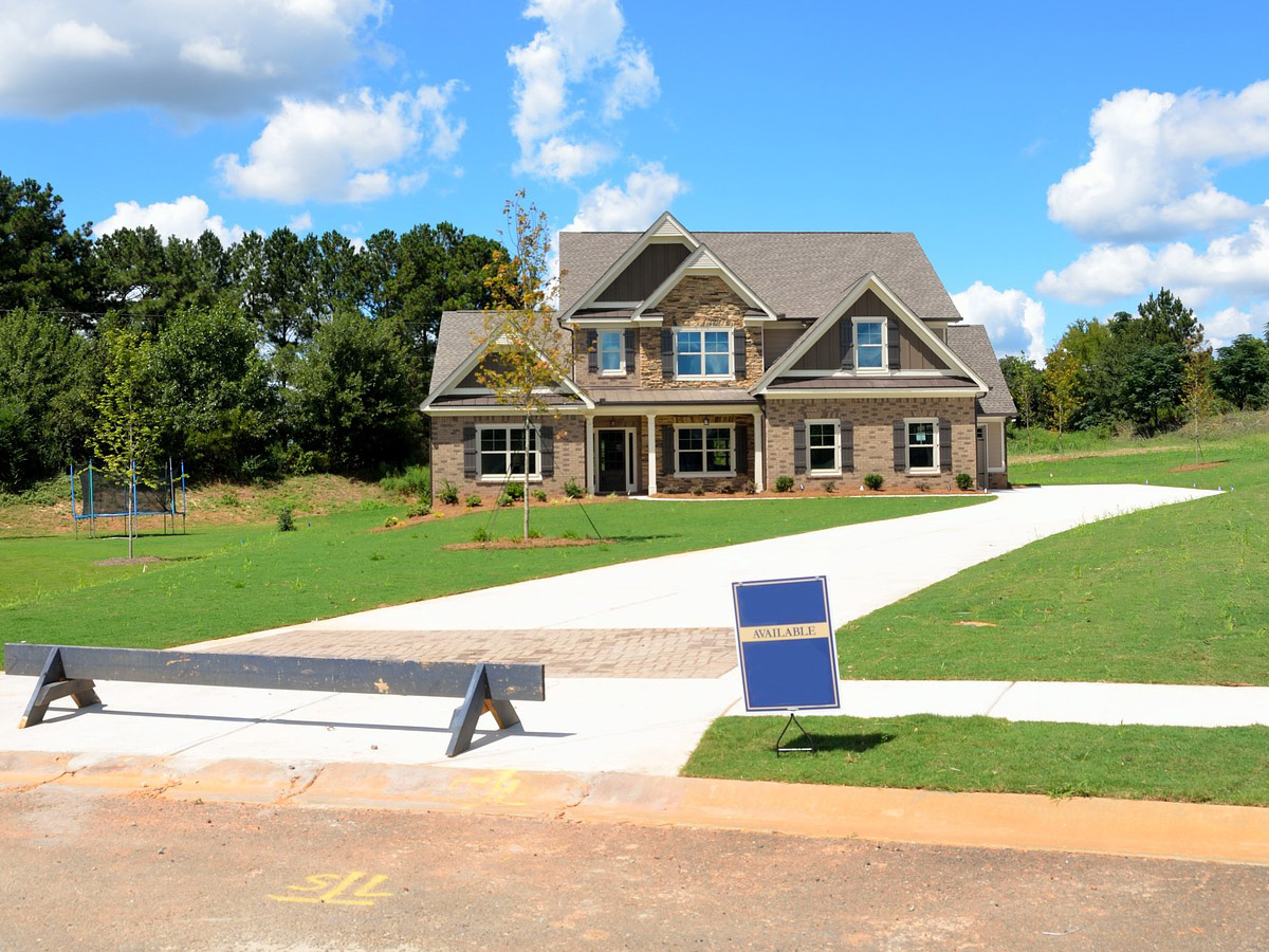 The image you uploaded features a two-story suburban house with a gabled roof and a mix of brick and siding façade. The house is set back on a large, lush green lawn with young trees and a clear blue sky above. A concrete driveway leads to the attached garage, and an "Available" sign is prominently displayed at the front, indicating the house is for sale. A wooden swing set is visible in the backyard, suggesting a family-friendly setting.